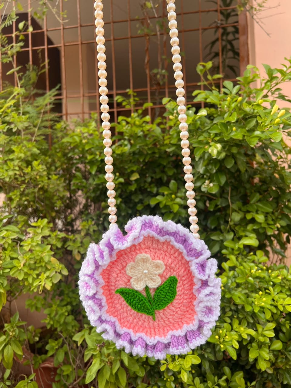 A round pink crochet bag with a cream flower, green leaves, lilac ruffles and a pearl-style beaded strap