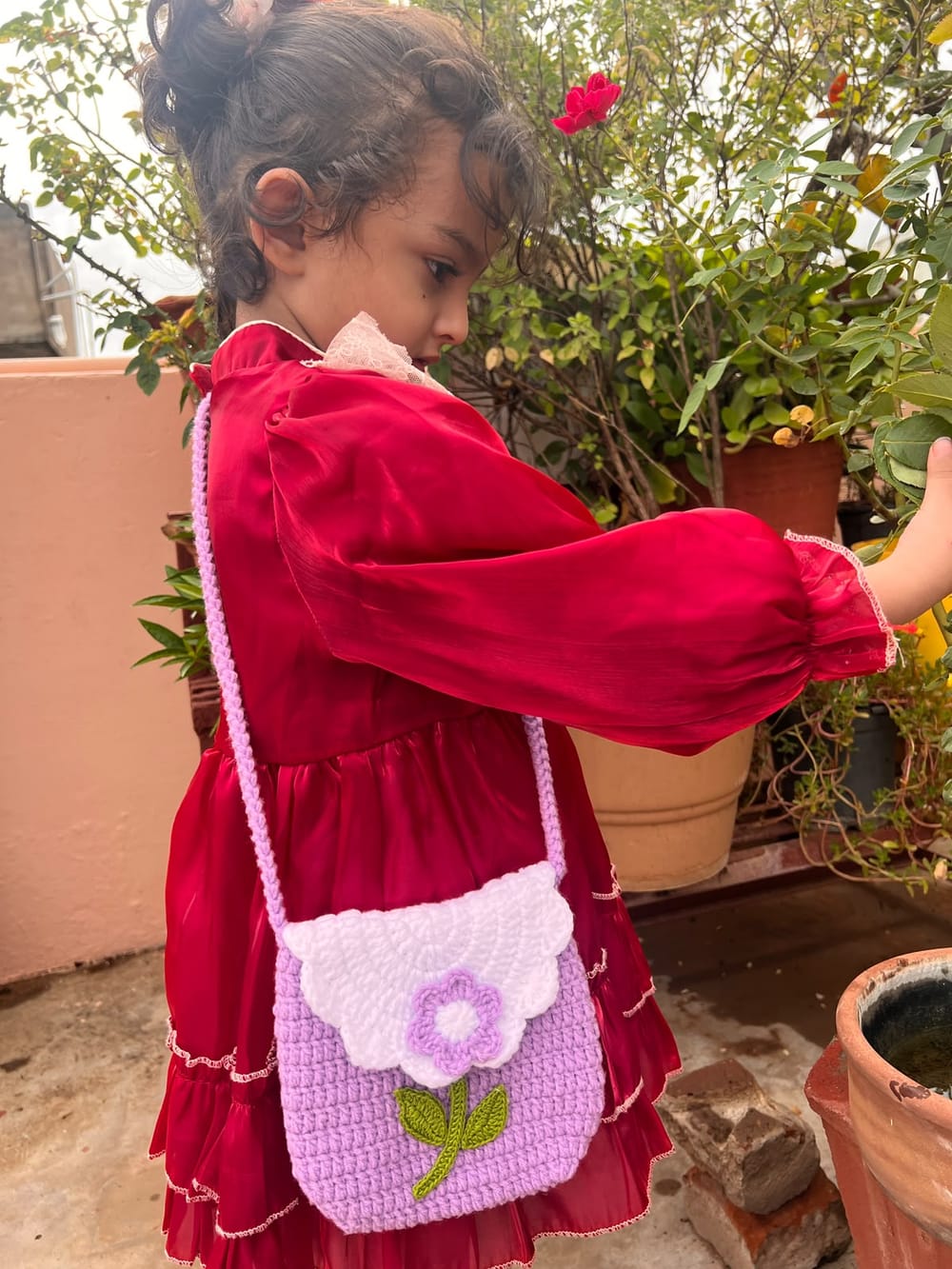 A child wearing a lilac crochet bag with a pale scalloped flap, purple flower and green leaves