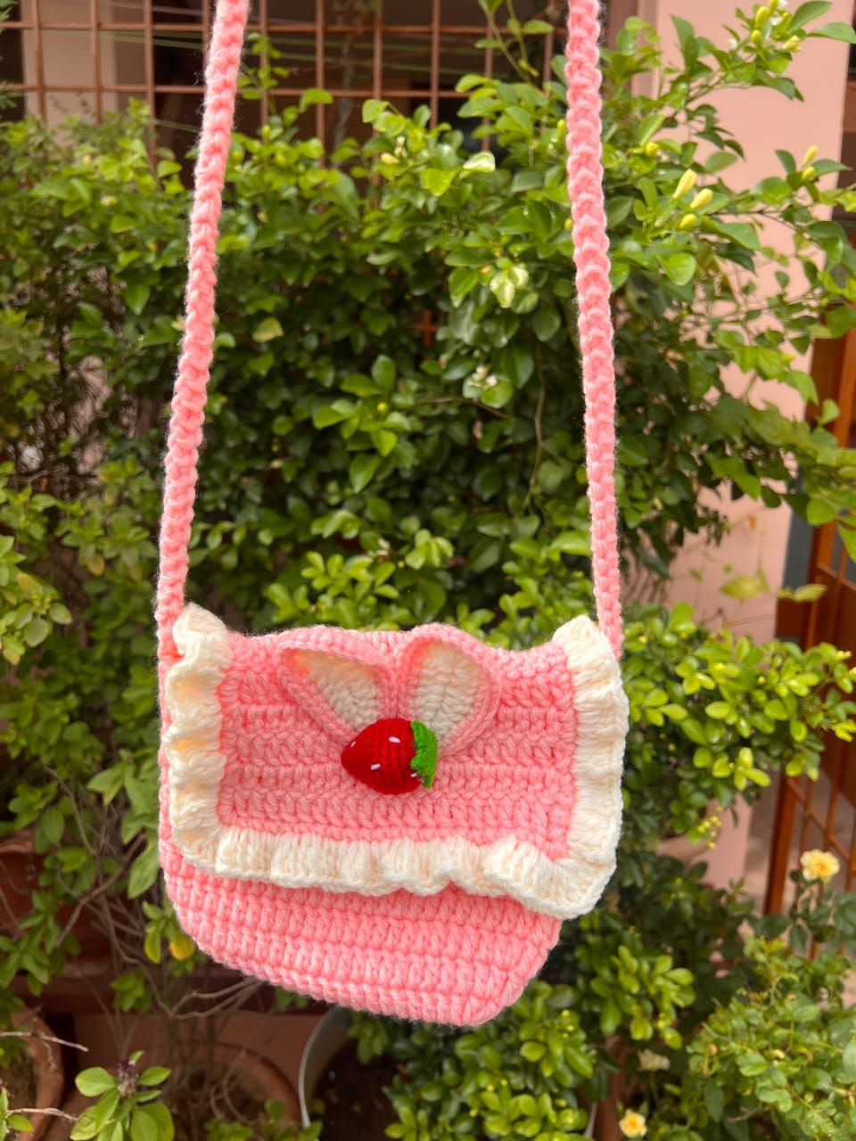 A pink crochet shoulder bag with cream ruffles, pale loops and a small red strawberry on the flap