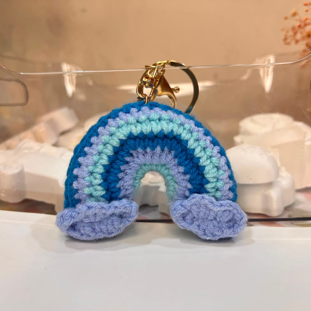 A hand-crocheted rainbow charm in teal, lilac and mint with lilac clouds, on a gold clip