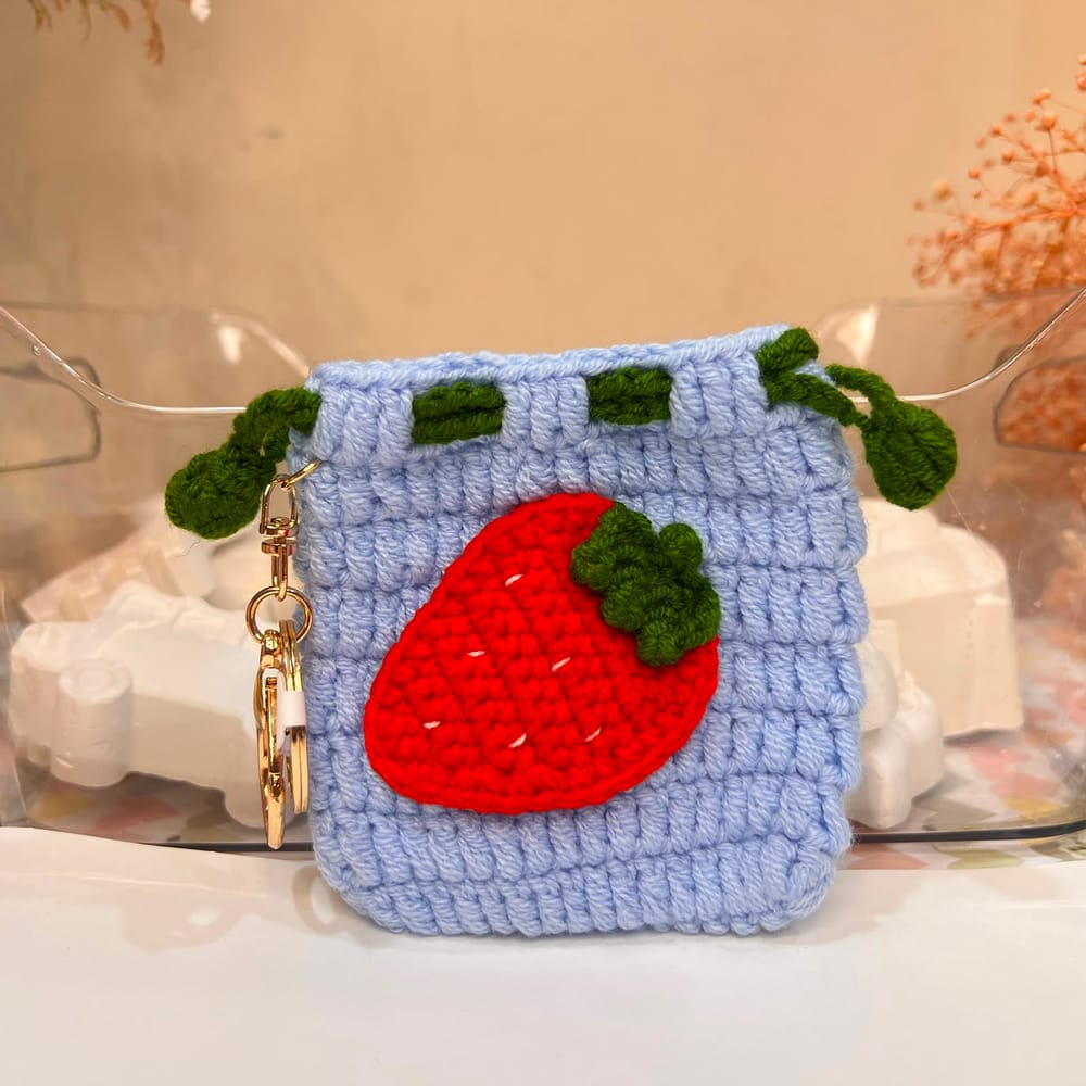A powder-blue hand-crocheted drawstring pouch with a large red strawberry, white seed stitches and green leaf loops