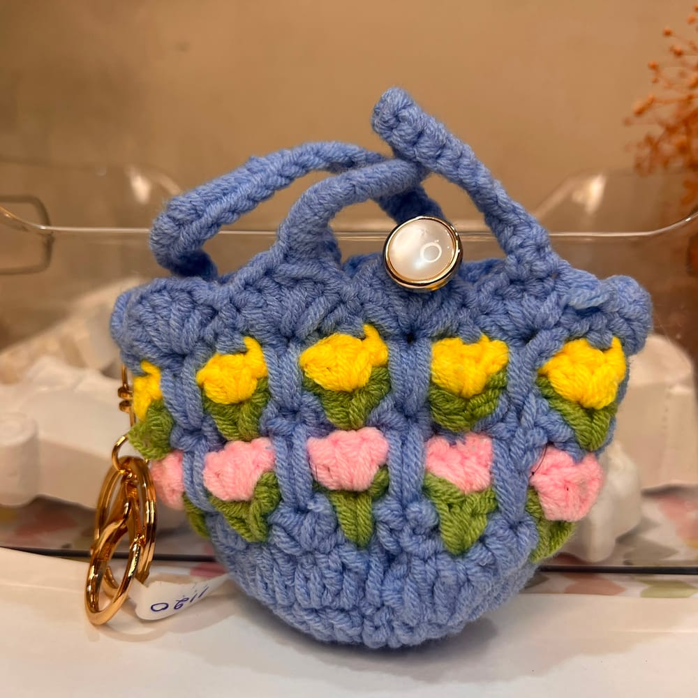 A cornflower blue hand-crocheted mini pouch with rows of yellow and pink tulips, a pearl button and a gold clip