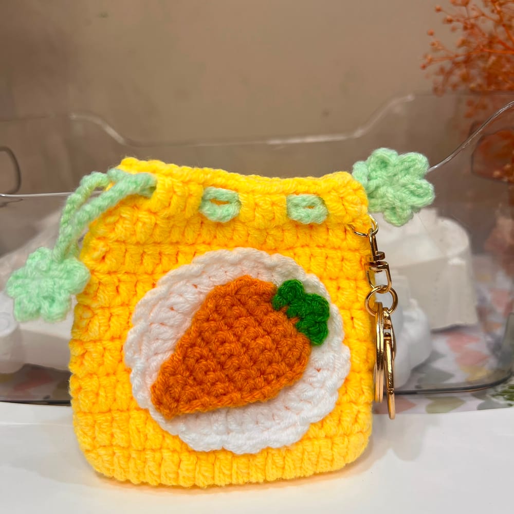 A yellow hand-crocheted drawstring pouch with an orange carrot on a white scalloped disc and mint-green flower drawstrings