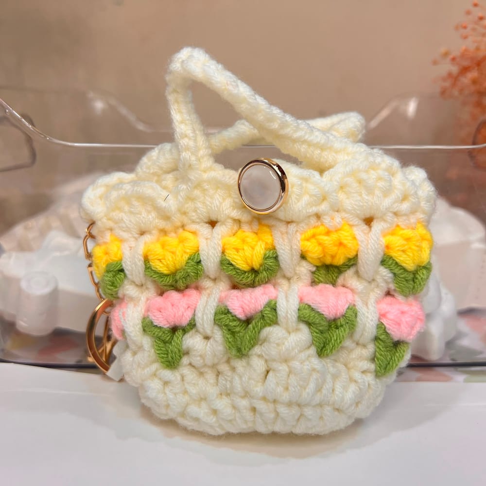 A cream hand-crocheted mini pouch with rows of yellow and pink tulips, a pearl button and a gold clip