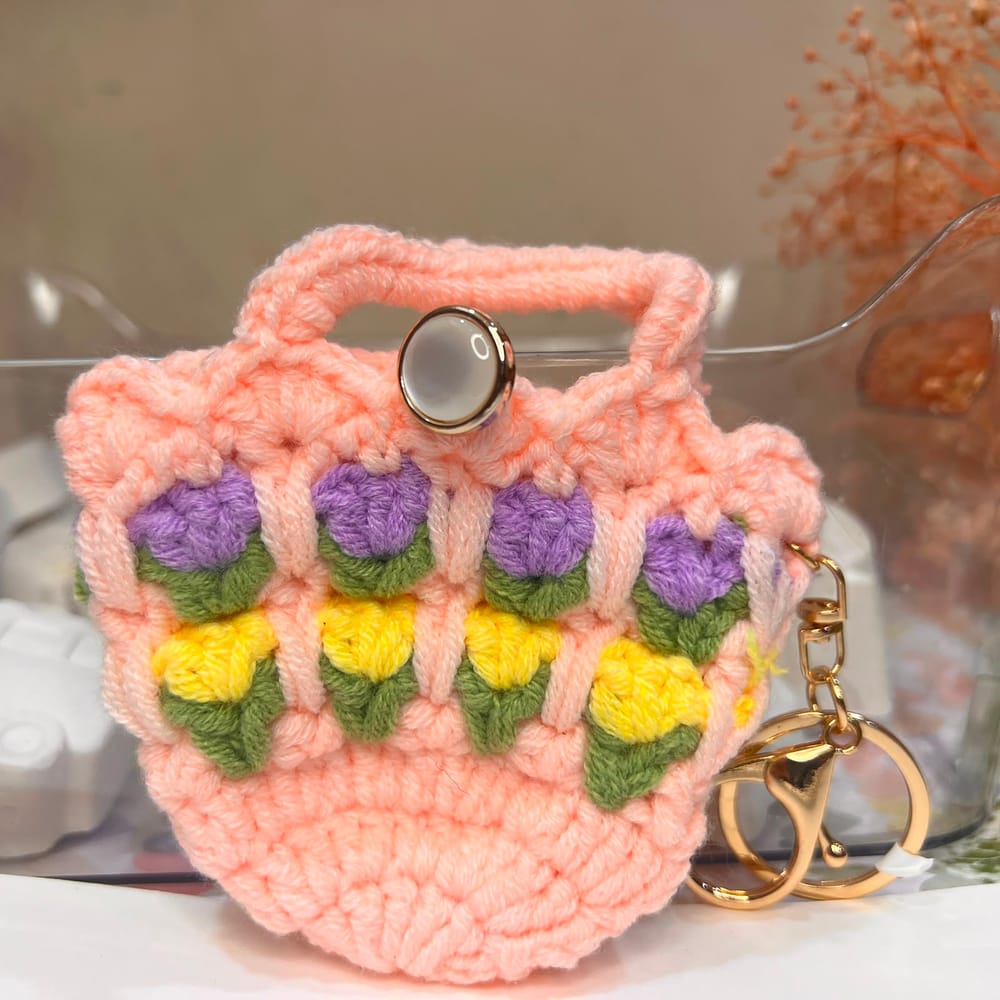 A pale peach hand-crocheted mini pouch with rows of purple and yellow tulips, a pearl button and a gold clip