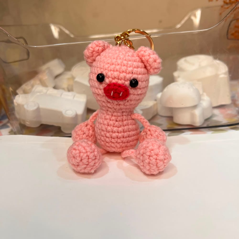 A pink hand-crocheted sitting piglet charm with a red snout and black bead eyes, on a gold clip