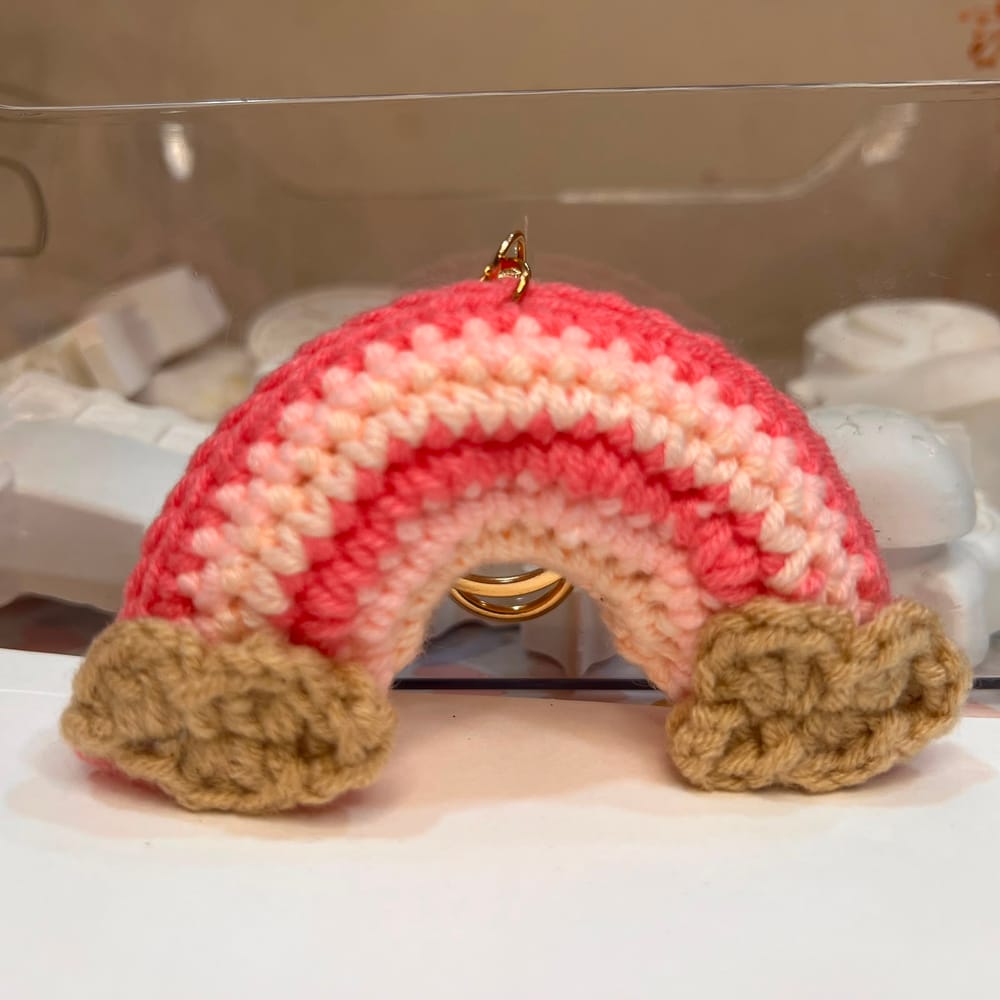 A hand-crocheted rainbow charm in coral, blush and cream with sand-coloured clouds, on a gold ring