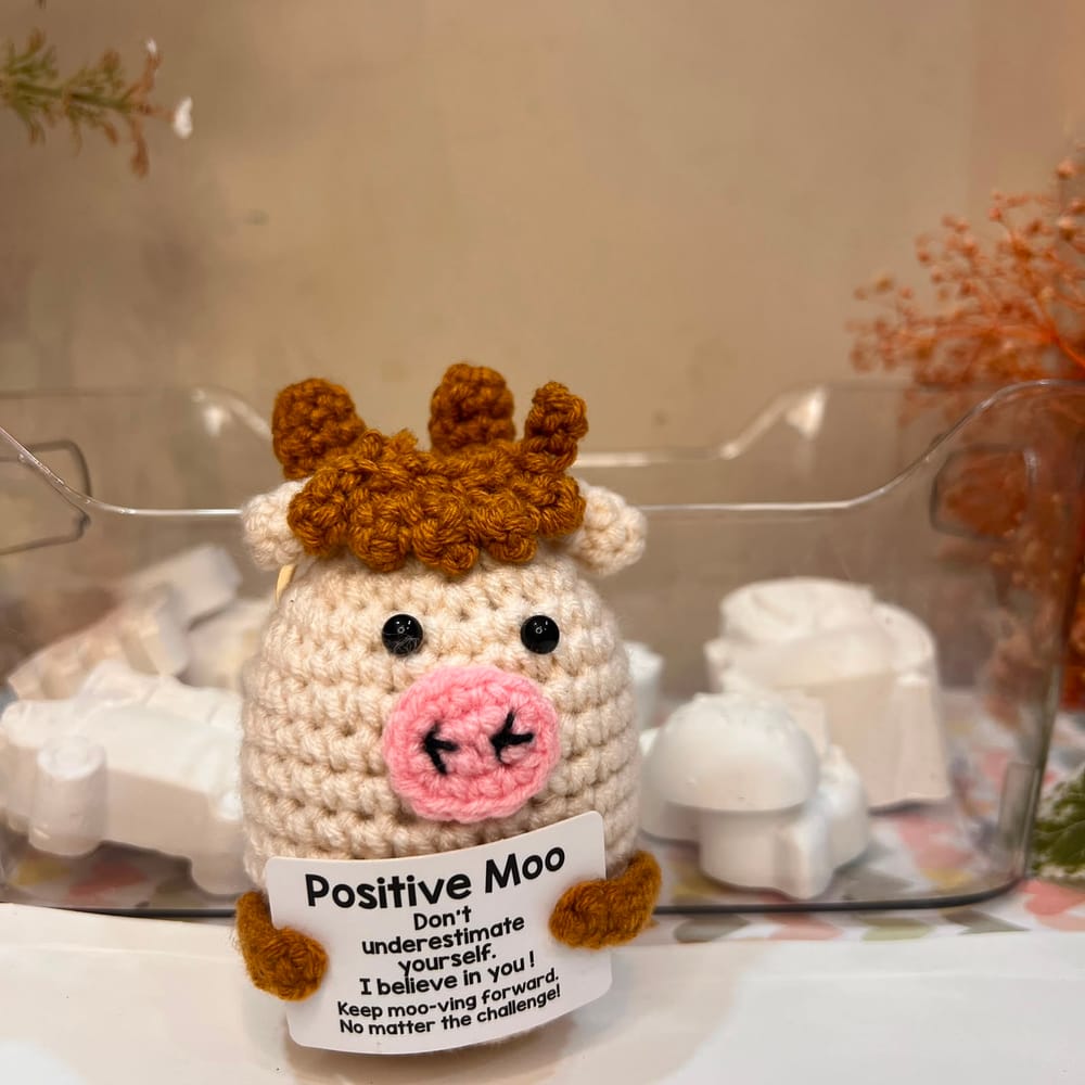 A cream hand-crocheted cow with a caramel fringe and pink snout, holding a card reading "Positive Moo, don't underestimate yourself, I believe in you"
