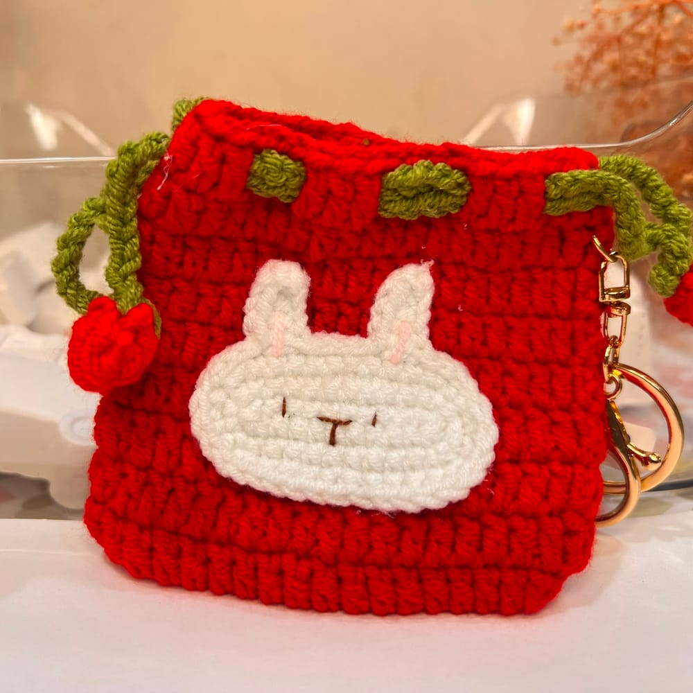 A red hand-crocheted drawstring pouch with a white sleepy bunny face, green leaf loops and a gold clip
