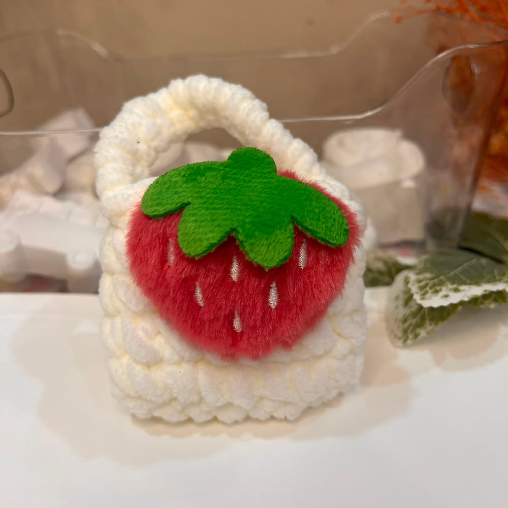 A white cloud-puff crochet pouch with a plush red strawberry and a green felt leaf on the front