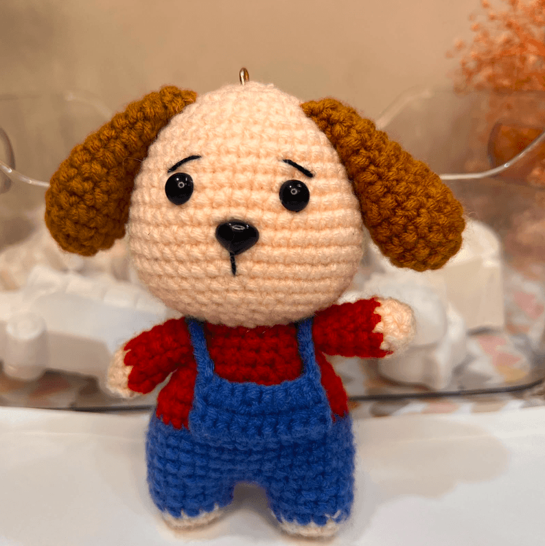 A hand-crocheted keychain toy with long floppy brown ears, wearing blue dungarees over a red top