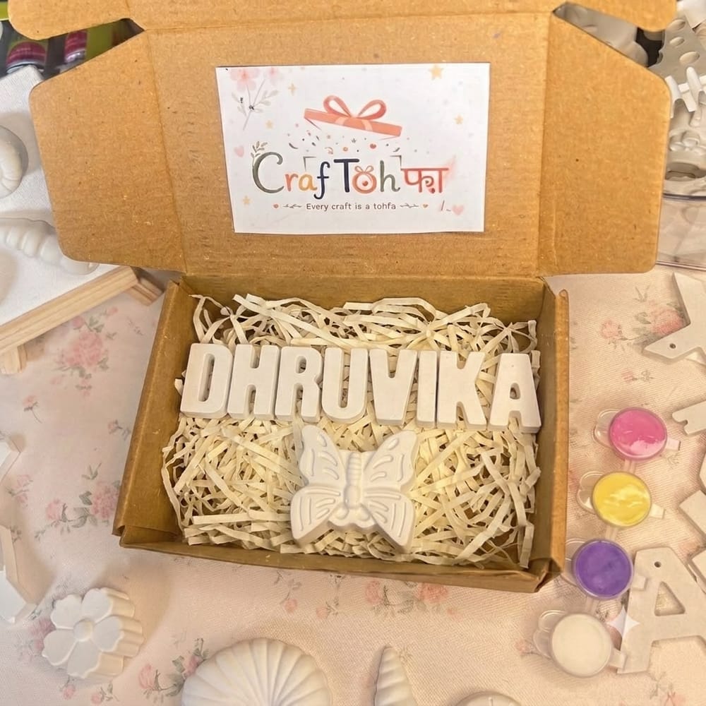 Unpainted plaster letters spelling "DHRUVIKA" with a plaster butterfly, paint pots and a brush, boxed on shredded paper