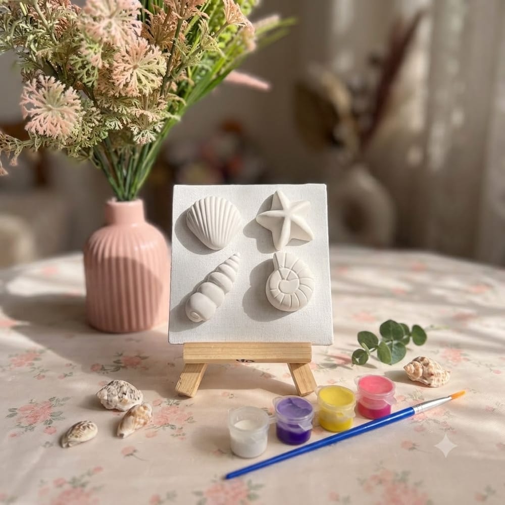 A small canvas on a wooden easel with an unpainted plaster scallop shell, starfish, spiral shell and ammonite mounted on it, beside four paint pots and a brush