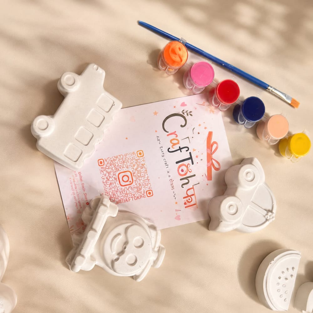 An unpainted plaster train engine, bus, panda face and watermelon slice with seven paint pots and a brush around a printed Craftohfa card